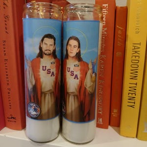 May include: Two glass candles featuring images of men in robes, halos, and USA shirts. The candles have a blue sky background and a white base. One man holds a medal. The candles are next to red and yellow books.