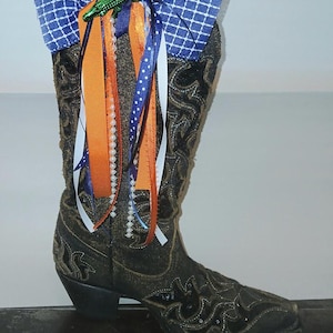 May include: A black cowboy boot featuring intricate dark stitching and a decorative arrangement at the top. The embellishment includes a blue and white checkered fabric bow, a green lizard figurine, and several hanging ribbons in orange, blue, and white.