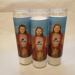 May include: Three cylindrical glass prayer candles featuring a graphic print of a person with long hair and a beard, wearing a red robe over a tan garment with a Colorado Avalanche hockey team logo on the chest, set against a blue sky background.