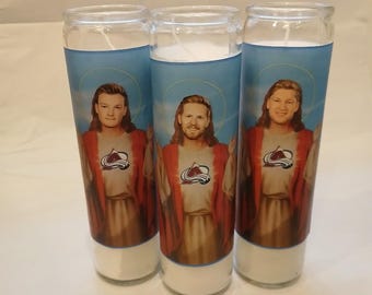 May include: Three glass prayer candles with a blue background featuring a portrait of a man in a red robe. The man is wearing a white shirt with a logo. The candles are white at the base and topped with a white wick.
