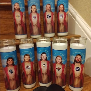 May include: Ten white candles with images of hockey players dressed as Jesus. Each candle features a different player and hockey team logo. The candles are arranged in two rows on a wooden surface. Two black hockey pucks are in the foreground.