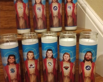 May include: Ten white candles with images of hockey players dressed as Jesus. Each candle features a different player and hockey team logo. The candles are arranged in two rows on a wooden surface. Two black hockey pucks are in the foreground.