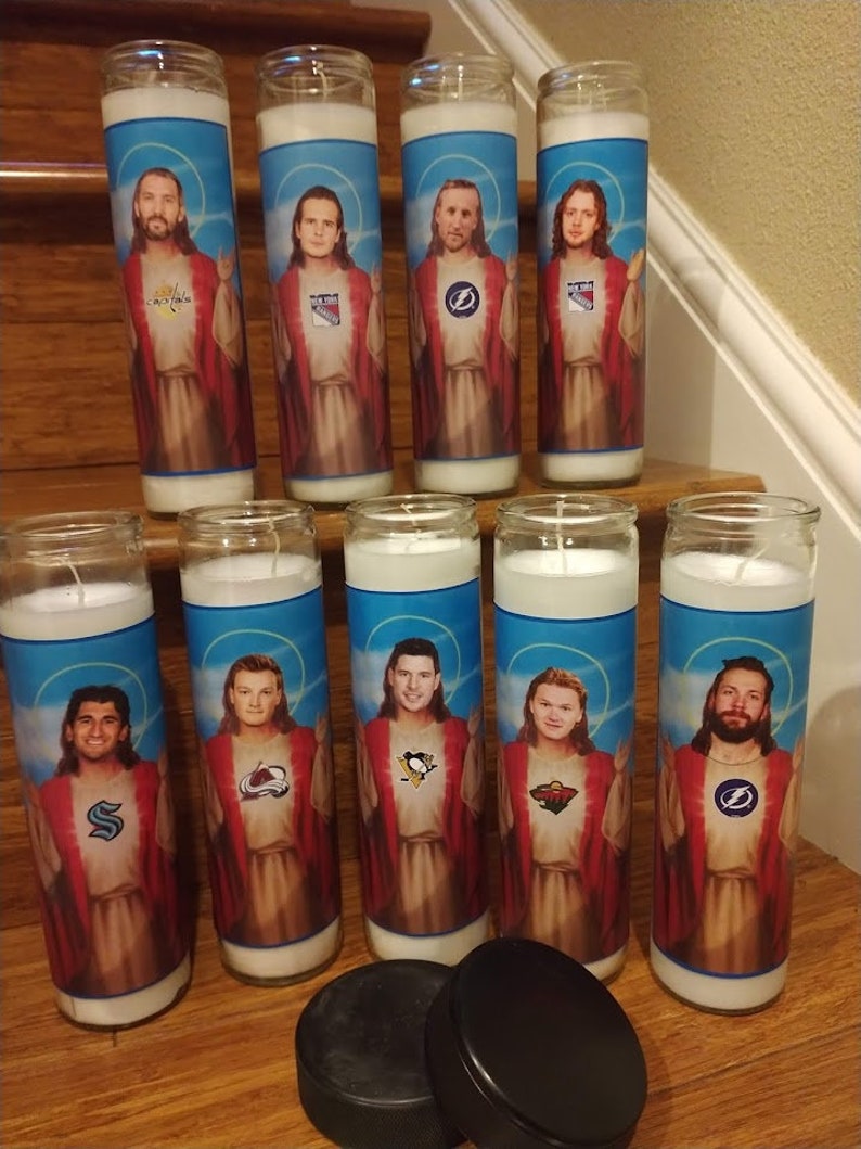 May include: Ten white candles with images of hockey players dressed as Jesus. Each candle features a different player and hockey team logo. The candles are arranged in two rows on a wooden surface. Two black hockey pucks are in the foreground.