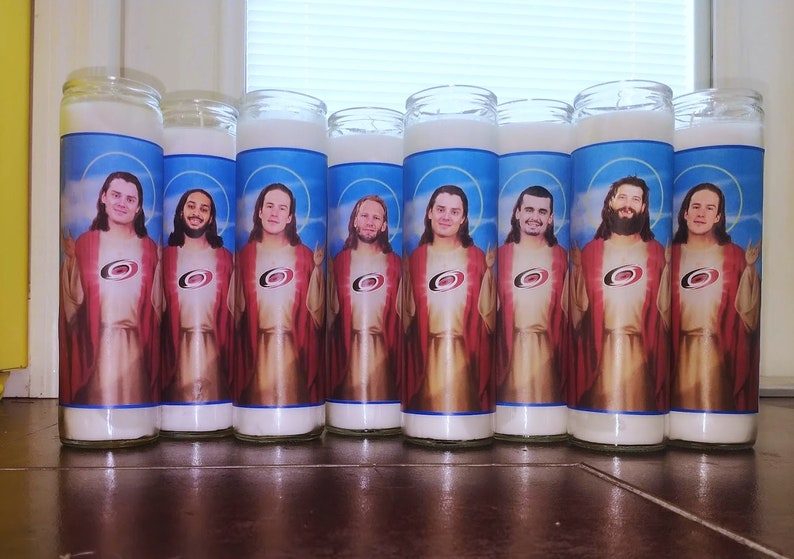 May include: Eight white candles with images of hockey players dressed as Jesus. Each candle features a different player wearing a red and black jersey with a white logo. The candles are arranged in a row on a wooden surface.