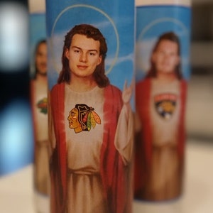 May include: Three white pillar candles with blue and red labels. The labels feature images of people with long hair wearing red robes and a hockey team logo. The labels also have a blue halo around the people's heads.