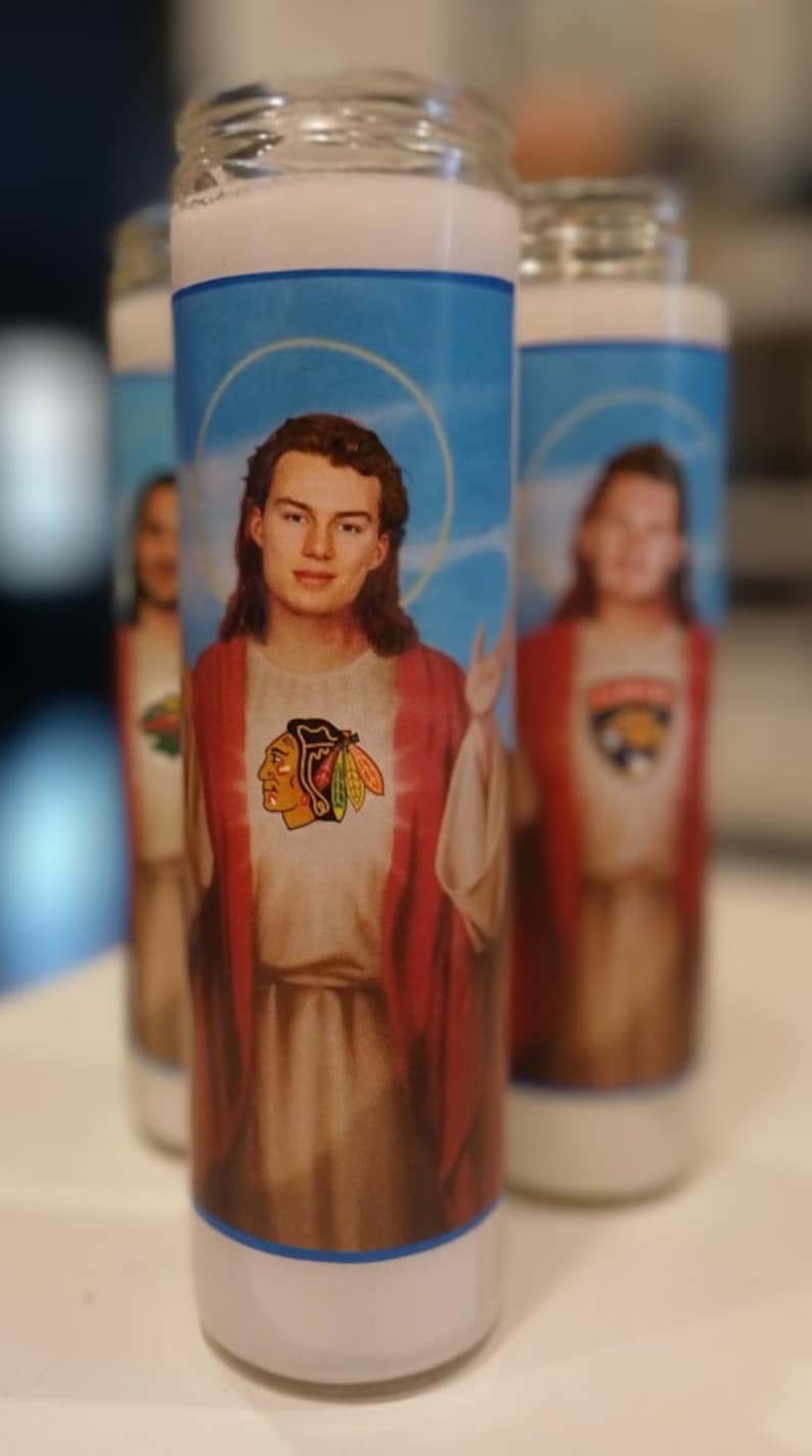 May include: Three white pillar candles with blue and red labels. The labels feature images of people with long hair wearing red robes and a hockey team logo. The labels also have a blue halo around the people's heads.