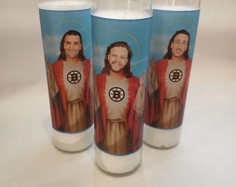 May include: Three tall cylindrical glass prayer candles filled with white wax. Each label features a figure in a red robe with a blue background and a yellow halo, wearing a white shirt with a black and yellow Boston Bruins hockey logo.