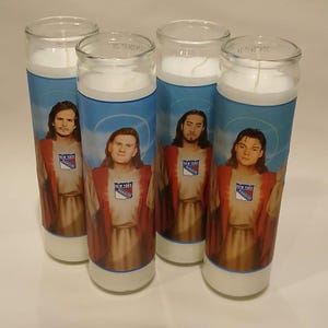 May include: Four cylindrical glass prayer candles filled with white wax. Each candle features a label depicting a person with long hair wearing a robe, with a New York Rangers logo on the chest and a light blue halo graphic behind the head.