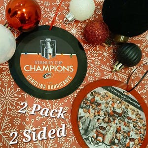 May include: Two round ornaments featuring Carolina Hurricanes Stanley Cup Champions 2026 branding. One ornament is black with orange text and a trophy graphic, the other is red with a team photo and trophy. Includes red hanging strings.
