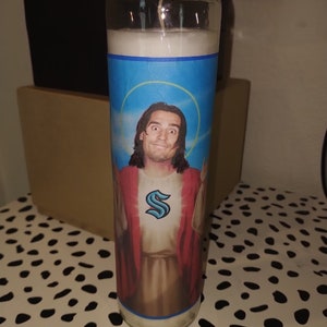 May include: A white candle with a blue, yellow, and white image of a man with long brown hair wearing a red robe with a blue "S" on the chest. The man is holding his arms out in a gesture of blessing.