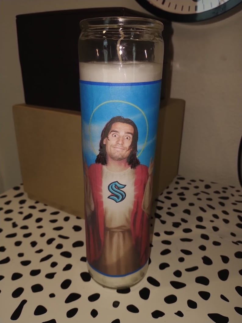 May include: A white candle with a blue, yellow, and white image of a man with long brown hair wearing a red robe with a blue "S" on the chest. The man is holding his arms out in a gesture of blessing.