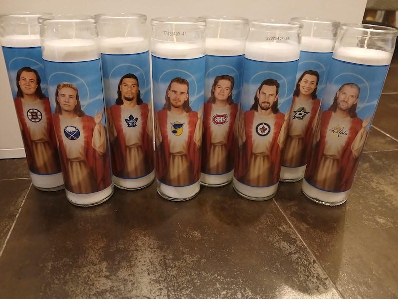 May include: Eight white candles with images of hockey players wearing jerseys of different teams. The players are depicted as Jesus Christ with halos and robes. The teams represented are the Boston Bruins, the Washington Capitals, the Tampa Bay Lightning, the Montreal Canadiens, the Toronto Maple Leafs, the Dallas Stars, the Philadelphia Flyers, and the Chicago Blackhawks.