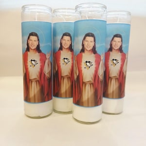 May include: Four tall cylindrical glass prayer candles, each featuring a label with a portrait of a man with long brown hair wearing a red robe over a tan garment, with a Pittsburgh Penguins hockey logo on the chest and a light blue sky background.