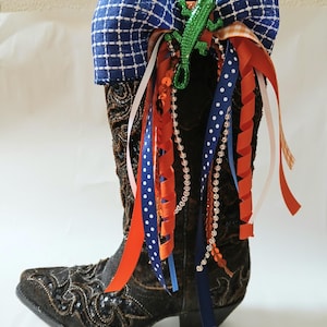 May include: A decorative brown cowboy boot adorned with a blue plaid bow, a green alligator, and colorful ribbons. The boot features intricate embroidery and is embellished with orange, white, and blue ribbons and beaded accents.
