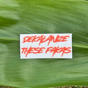 May include: A white sticker with the red text "DEKĀLĀNIZE THESE FĀKĀS" is displayed on a green leaf. The sticker is rectangular and the text is in a bold, stylized font. The background is a green leaf and grass.