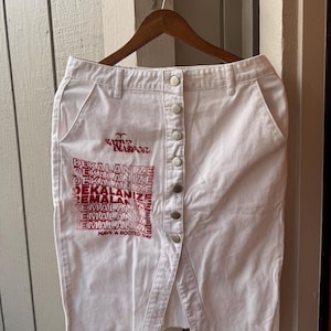 May include: A white denim skirt featuring a front button closure and a raw, frayed hem. The fabric is printed with red text that reads DEKALANIZE and HAVE A ROOTED DAY, along with a small graphic symbol above the repeated text.