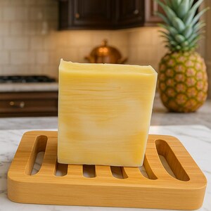 May include: A large, rectangular bar of yellow soap sits on a bamboo soap dish. The soap has a marbled effect. A whole pineapple is visible in the background, suggesting a tropical theme. The soap dish has a slotted design.