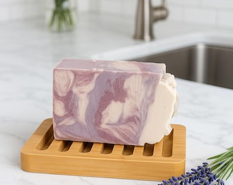 May include: A rectangular bar of lavender-scented soap with a marbled purple and white design rests on a bamboo soap dish. A sprig of lavender and a vase of lavender flowers are in the background. The soap dish has a slotted design.