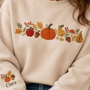 May include: A beige crewneck sweatshirt featuring an embroidered horizontal band of autumn motifs including a pumpkin, pear, apple, mushroom, acorns, and leaves. The left sleeve displays a smaller matching embroidery with the name Clara below it.