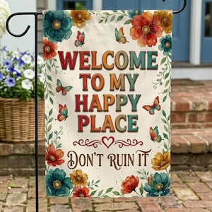 May include: A garden flag with the text "WELCOME TO MY HAPPY PLACE DON'T RUIN IT" in colorful letters. The flag is decorated with floral and butterfly designs in shades of red, teal, and yellow. The flag is displayed on a black metal stand.