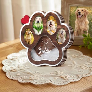 May include: A wooden paw-shaped photo frame with five openings for pictures of dogs. The frame has a brown center and a white border. The name "Buddie" and the date "24.12.2025" are displayed. A small red heart is attached to the frame.