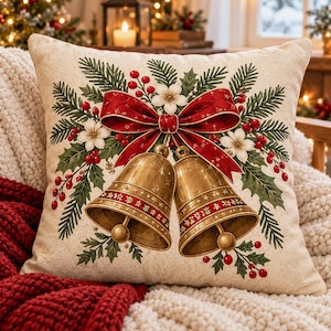 May include: A square throw pillow with a cream-colored background featuring a central illustration of two gold bells tied with a large red ribbon. The design includes green pine branches, holly leaves, red berries, and small white flowers.