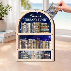 May include: Wooden bookshelf-shaped money bank with the text "Emma's LIBRARY FUND". The bank features three shelves filled with decorative book images. A hand is inserting a $100 bill into the slot at the top. The background shows a blue starry sky.