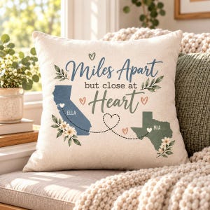 May include: A square throw pillow with a cream-colored cover featuring the text Miles Apart but close at Heart. The design includes a blue map of California labeled Ella and a green map of Texas labeled Mia, connected by a dashed heart line.