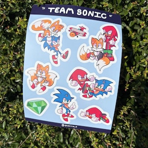 May include: A sheet of stickers featuring characters from the Sonic the Hedgehog franchise. The stickers include Sonic, Tails, Knuckles, and a green emerald, with the text "= TEAM SONIC =" at the top. The stickers are on a blue background.