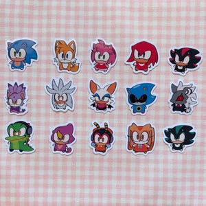 Puede incluir: Una colección de pegatinas de personajes de dibujos animados coloridos, con varios diseños de Sonic the Hedgehog y sus amigos. Las pegatinas están sobre un fondo a cuadros rosa y blanco. Cada pegatina tiene un borde blanco.