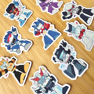 May include: A collection of nine die-cut stickers featuring stylised robot characters in various poses. The robots are depicted in vibrant colours including blue, red, yellow, purple, white, and black, each with distinct mechanical features and expressions.