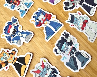 May include: A collection of nine die-cut stickers featuring stylised robot characters in various poses. The robots are depicted in vibrant colours including blue, red, yellow, purple, white, and black, each with distinct mechanical features and expressions.