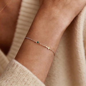 May include: Gold-toned bracelet featuring a delicate chain, a small green gemstone, and a cross charm. The bracelet is shown on a wrist, suggesting a piece of jewelry designed for everyday wear or special occasions.
