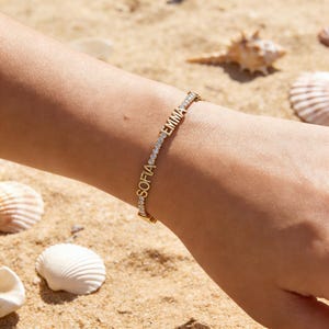 May include: A gold bracelet with the names "SOFIA" and "EMMA" spelled out in gold letters. The bracelet is adorned with small, sparkling stones. The bracelet is on a wrist, with seashells and sand in the background.