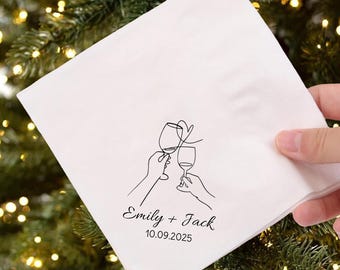May include: White napkin with a black line drawing of two hands toasting wine glasses. The names "Emily + Jack" and the date "10.09.2025" are printed below the illustration. The napkin is held against a background of Christmas tree branches.
