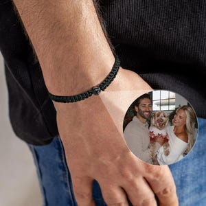 Peut inclure: Bracelet noir tressé avec une perle ronde foncée. Le bracelet est porté au poignet d'une personne. Une petite image circulaire montre un couple avec un chien.