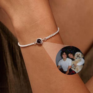 May include: A silver bracelet with a small, round, dark purple stone in the center. The bracelet has a delicate, textured chain design. A small, circular image shows a couple and a dog.