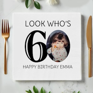 Puede incluir: Una servilleta cuadrada blanca con el texto "LOOK WHO'S 60" y "HAPPY BIRTHDAY EMMA" en negro. Una foto de una niña pequeña está en el centro. Un tenedor y un cuchillo dorados están a los lados.