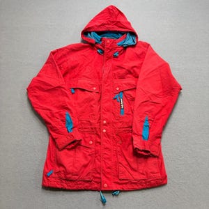 May include: A red hooded jacket with a blue interior and accents. The jacket features multiple pockets, a zipper closure, and a drawstring at the hem. The jacket has the brand name "Helly Hansen" on the front.