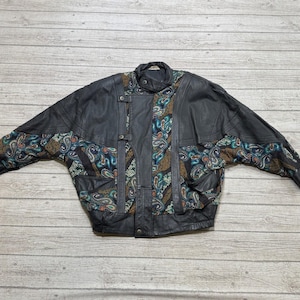 May include: A black leather jacket featuring panels of multicolored paisley patterned fabric on the sleeves and front. The jacket includes a stand-up collar, a front button closure, and long sleeves with a wide, relaxed fit design.