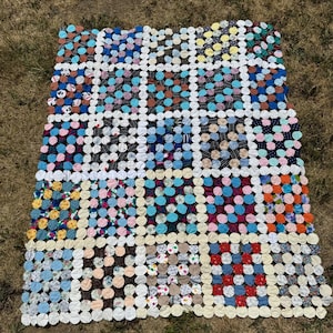 Peut inclure: Une courtepointe faite à la main composée de nombreuses petites pièces de tissu circulaires dans une grande variété de couleurs et de motifs, dont le bleu, le rouge, le jaune et le noir, reliées par des connecteurs en tissu blanc en grille.