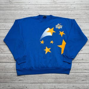 May include: A royal blue long-sleeve sweatshirt featuring a graphic print of yellow stars and a silver comet tail. The design includes the text "IMAGINE" in white lettering near the top right, accompanied by smaller graphic elements above the word.