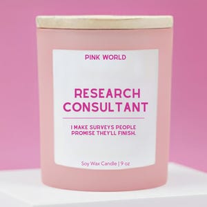 May include: A pink soy wax candle with a wooden lid. The label reads "PINK WORLD RESEARCH CONSULTANT I MAKE SURVEYS PEOPLE PROMISE THEY'LL FINISH." The candle is 9 oz.