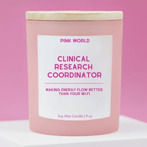 May include: A pink soy wax candle with a wooden lid. The label reads "PINK WORLD CLINICAL RESEARCH COORDINATOR MAKING ENERGY FLOW BETTER THAN YOUR WI-FI." The candle is 9 oz.