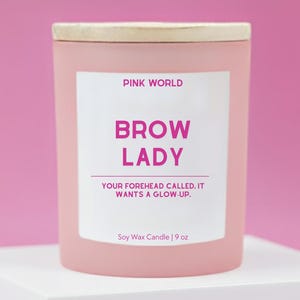 May include: A pink soy wax candle with a wooden lid. The label reads "PINK WORLD" and "BROW LADY" with the text "YOUR FOREHEAD CALLED. IT WANTS A GLOW-UP." The candle is 9 oz.