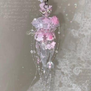 May include: A translucent pink and white jellyfish-shaped charm featuring delicate butterfly accents, small floral embellishments, and dangling beaded strands. The piece includes a silver-toned metal clasp at the top and the text Fragilebaby printed in white script across the center of the jellyfish body.