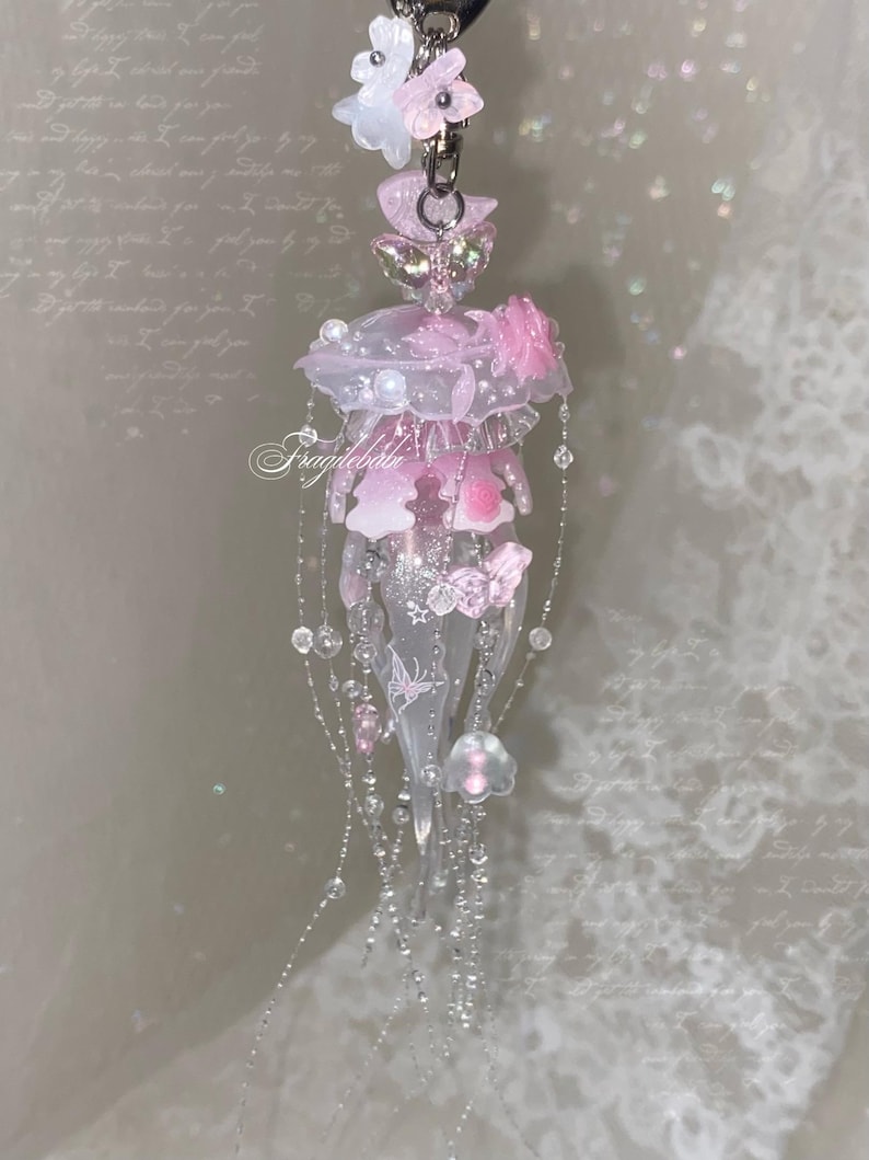 May include: A translucent pink and white jellyfish-shaped charm featuring delicate butterfly accents, small floral embellishments, and dangling beaded strands. The piece includes a silver-toned metal clasp at the top and the text Fragilebaby printed in white script across the center of the jellyfish body.