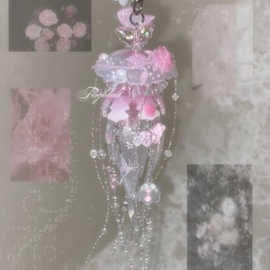 May include: A translucent pink and white jellyfish-shaped charm featuring delicate floral accents, small beads, and dangling strands. The piece includes a metal clasp at the top and displays the text Fragilebals in a cursive script across the center.