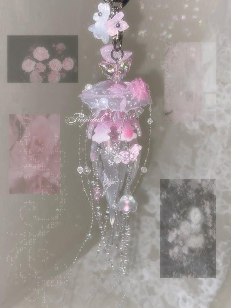 May include: A translucent pink and white jellyfish-shaped charm featuring delicate floral accents, small beads, and dangling strands. The piece includes a metal clasp at the top and displays the text Fragilebals in a cursive script across the center.