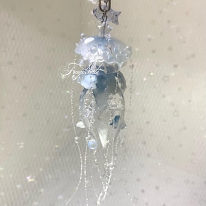 May include: A clear and blue jellyfish-shaped keychain with a silver clasp. The jellyfish has a translucent body with delicate details and dangling beaded strands. The keychain is adorned with small flowers and stars.